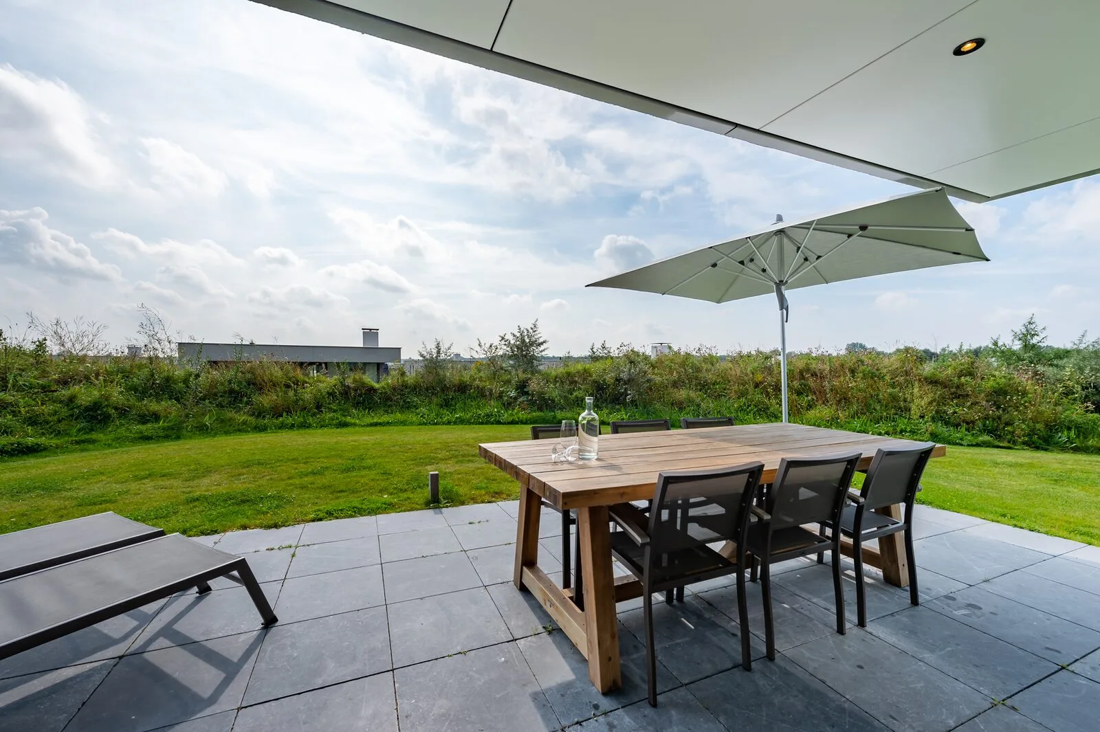 A paved outdoor patio has a wooden dining table and chairs beneath a large umbrella, overlooking a green lawn.