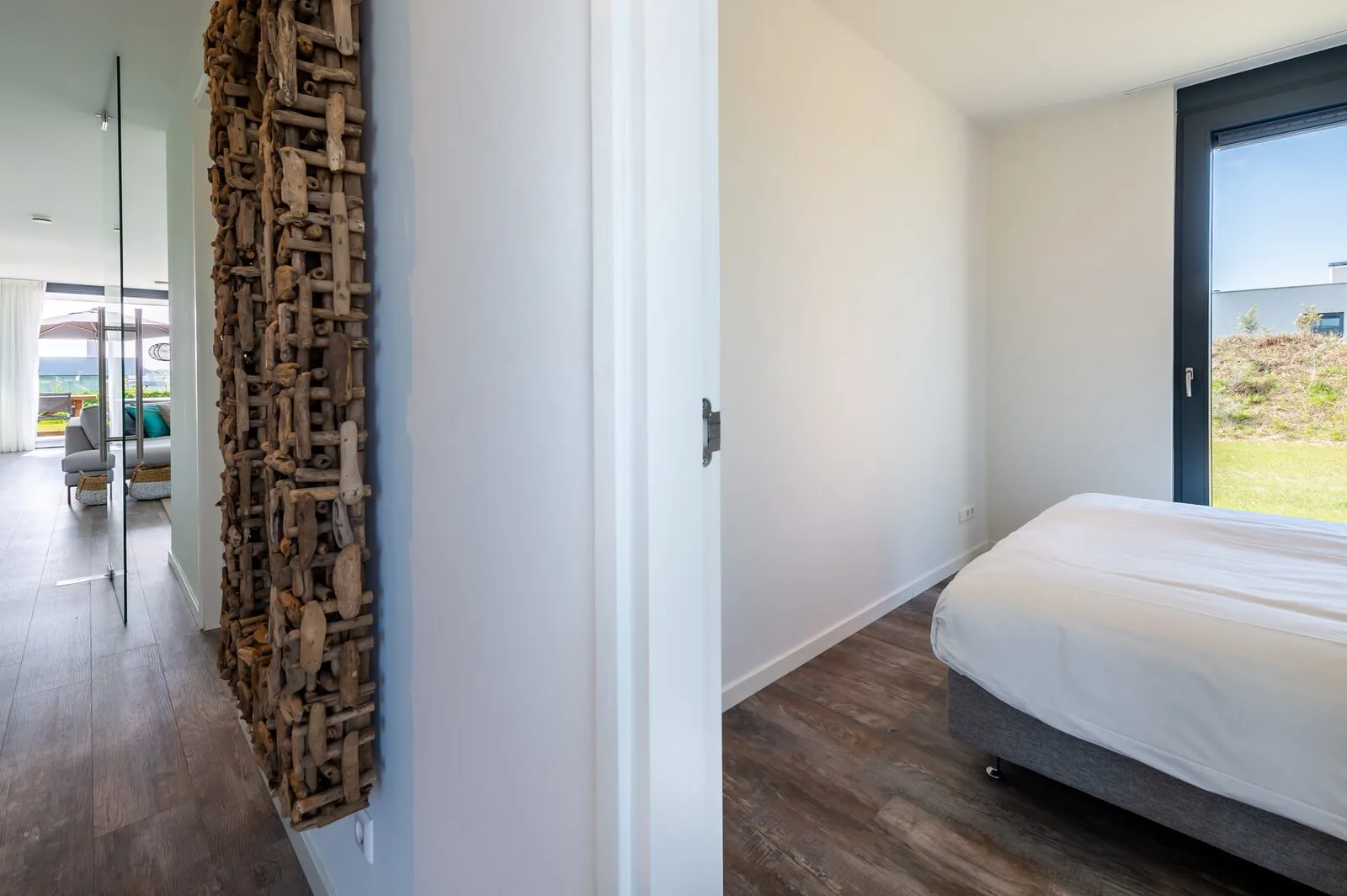 A bright bedroom has a white bed beside a large glass door overlooking greenery.