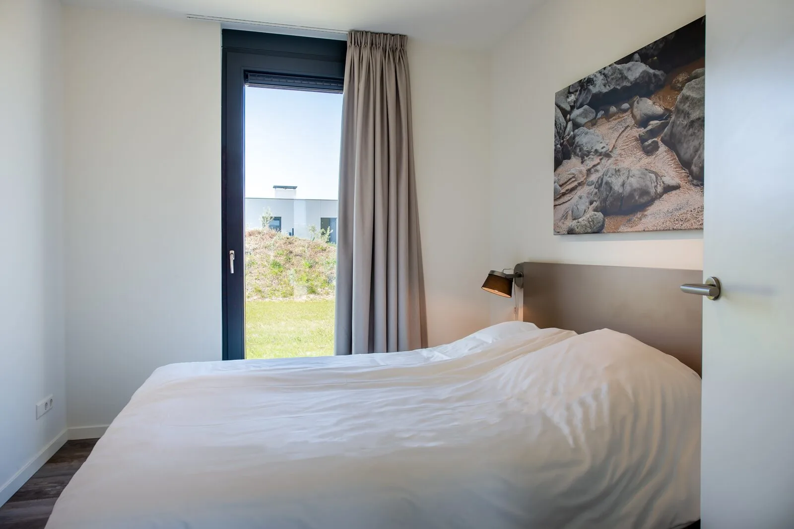 A bedroom with a double bed, gray headboard, bedside lamp, and glass door opening to a garden.