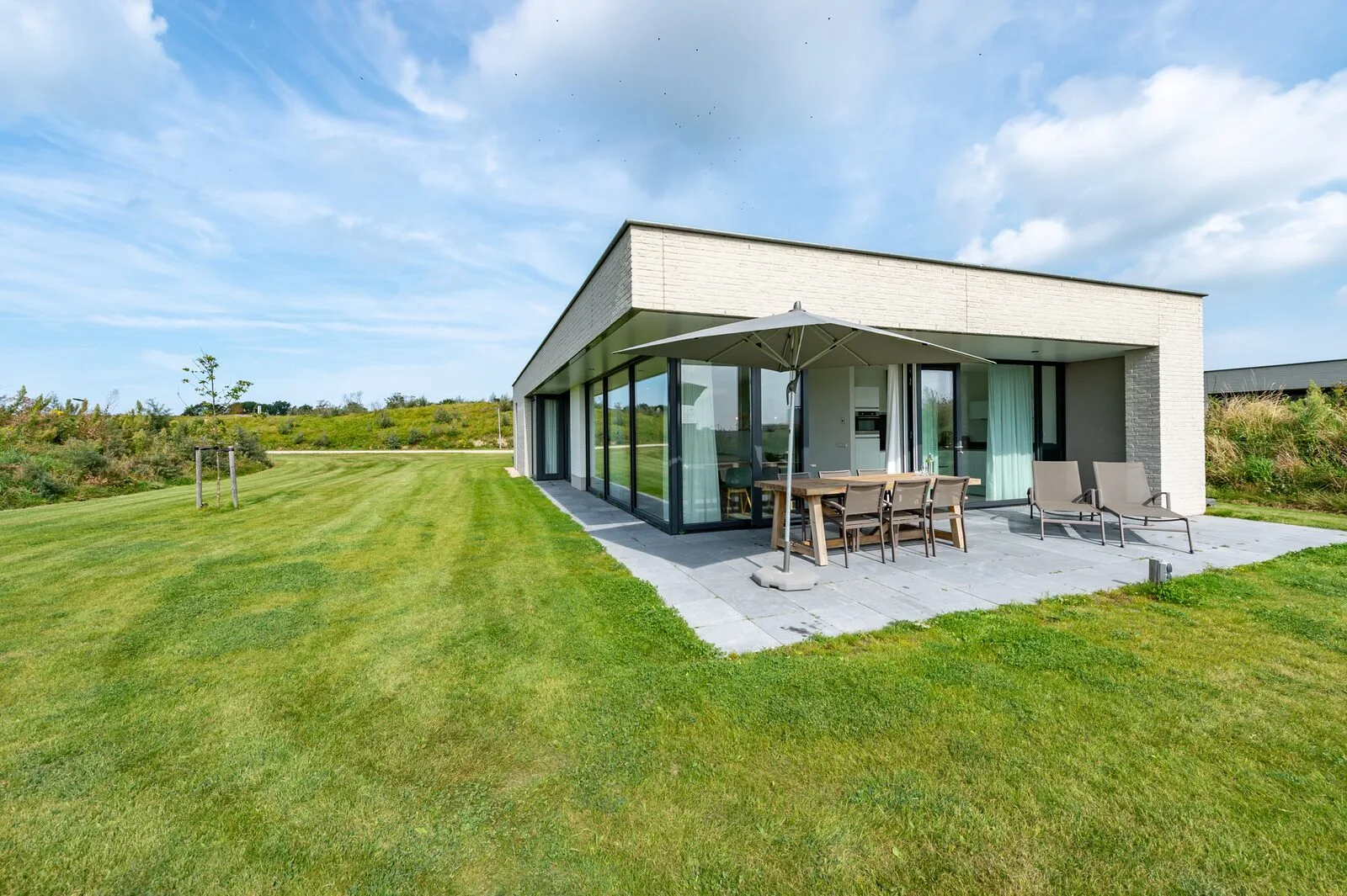 Modern single-story house with large glass walls, a paved patio, outdoor dining table, and lawn.