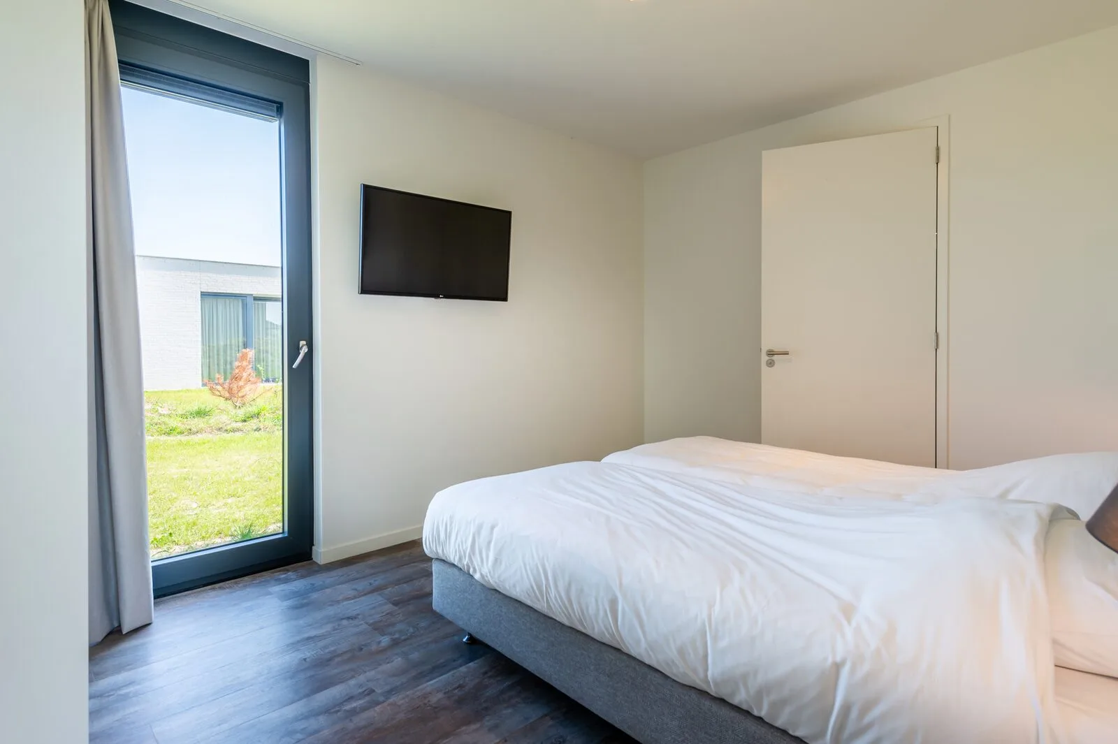 A bedroom with a white bed, wall-mounted television, and a tall window overlooking a grassy area.