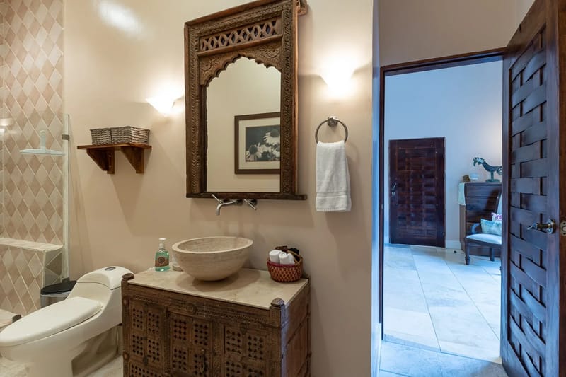 Villa Michelina · 20. This Balinese-inspired bathroom features a stone vessel sink set on a carved wooden vanity with a large matching mirror, alongside a tiled shower, and an open doorway leading to another area of the villa.