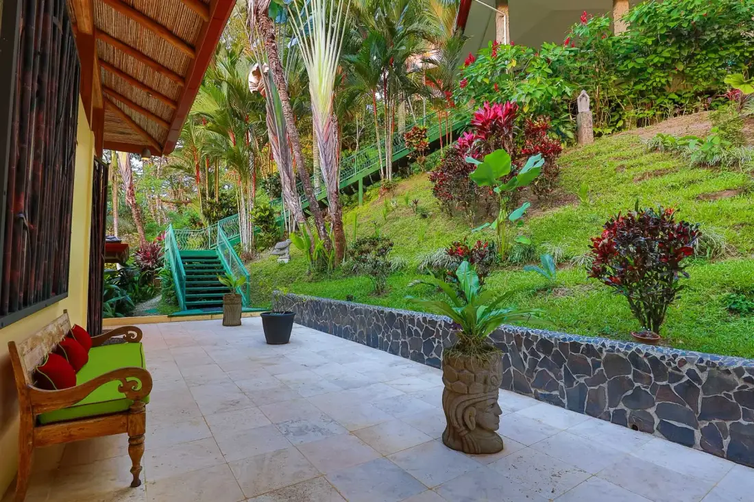 Una terraza exterior cubierta se abre a un jardín en una ladera con escalones verdes.