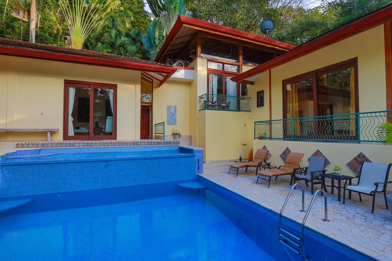 Villa Michelina · 23. The villa's yellow exterior, featuring distinctive red-tiled roofs and multiple balconies, overlooks a two-tier blue mosaic pool and a spacious stone patio surrounded by vibrant tropical foliage.