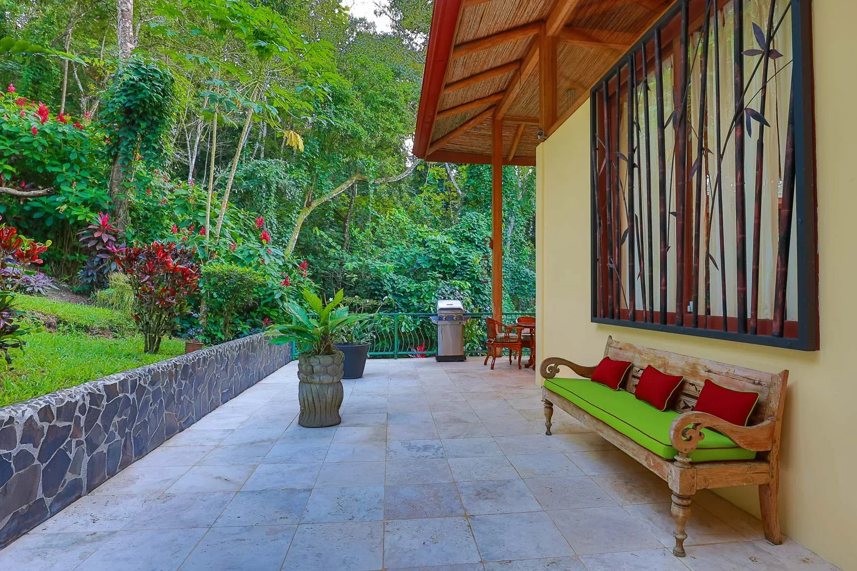 Un patio exterior cubierto con suelo de baldosas y asientos junto a un jardín frondoso.