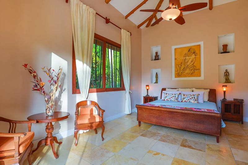 Villa Michelina · 17. A spacious Balinese-inspired bedroom features a king-size bed, a vaulted bamboo ceiling, and abundant natural light entering through a large window with views of lush greenery.