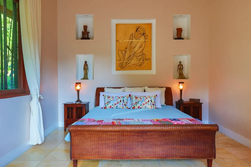 Villa Michelina · 16. A bedroom features a woven bed frame, built-in wall niches with artwork, and a window providing natural light and a view of lush greenery.