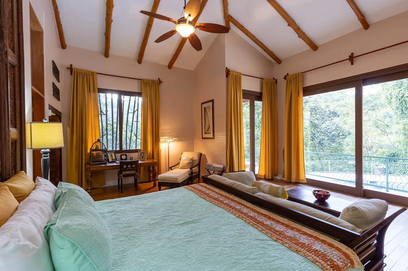 Villa Michelina · 12. A spacious bedroom with a king bed, vaulted ceiling, and exposed wood beams features ample natural light from large sliding glass doors that open to a balcony overlooking a tropical canopy.