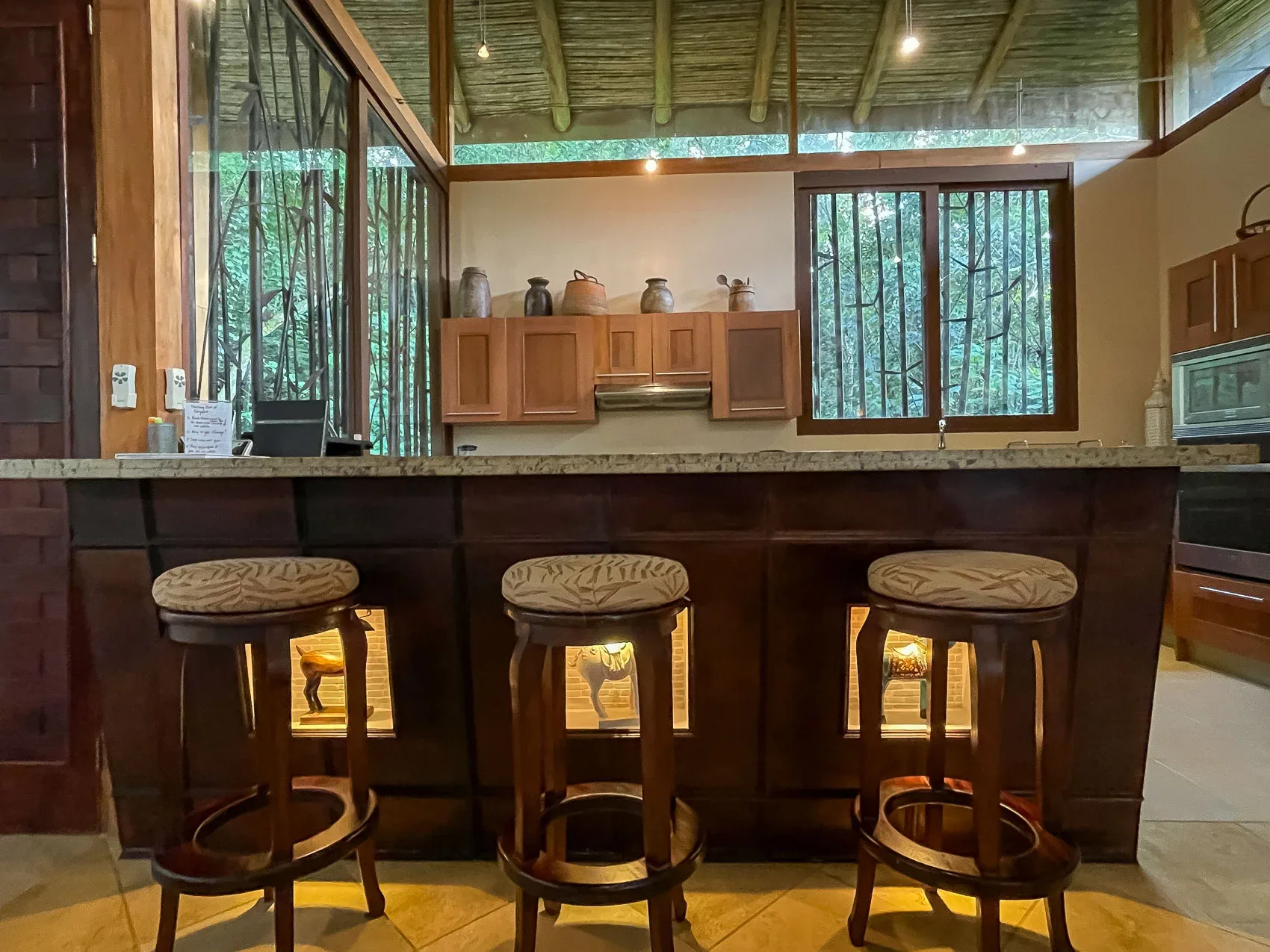 Una barra de cocina con taburetes está bajo un techo alto, junto a ventanas con vistas a vegetación frondosa.