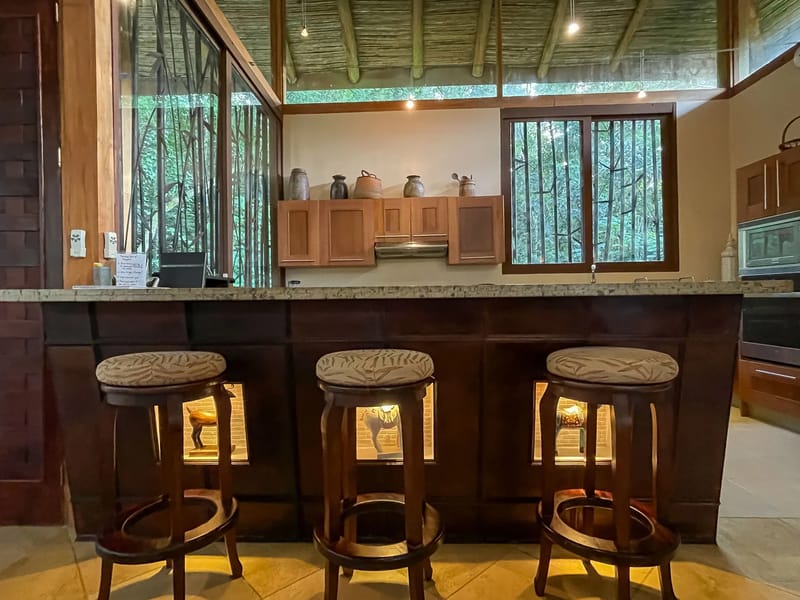 Villa Michelina · 10. A Balinese-inspired kitchen bar with three stools and wooden cabinetry features raised bamboo ceilings and large windows that provide natural light and views of dense tropical foliage.