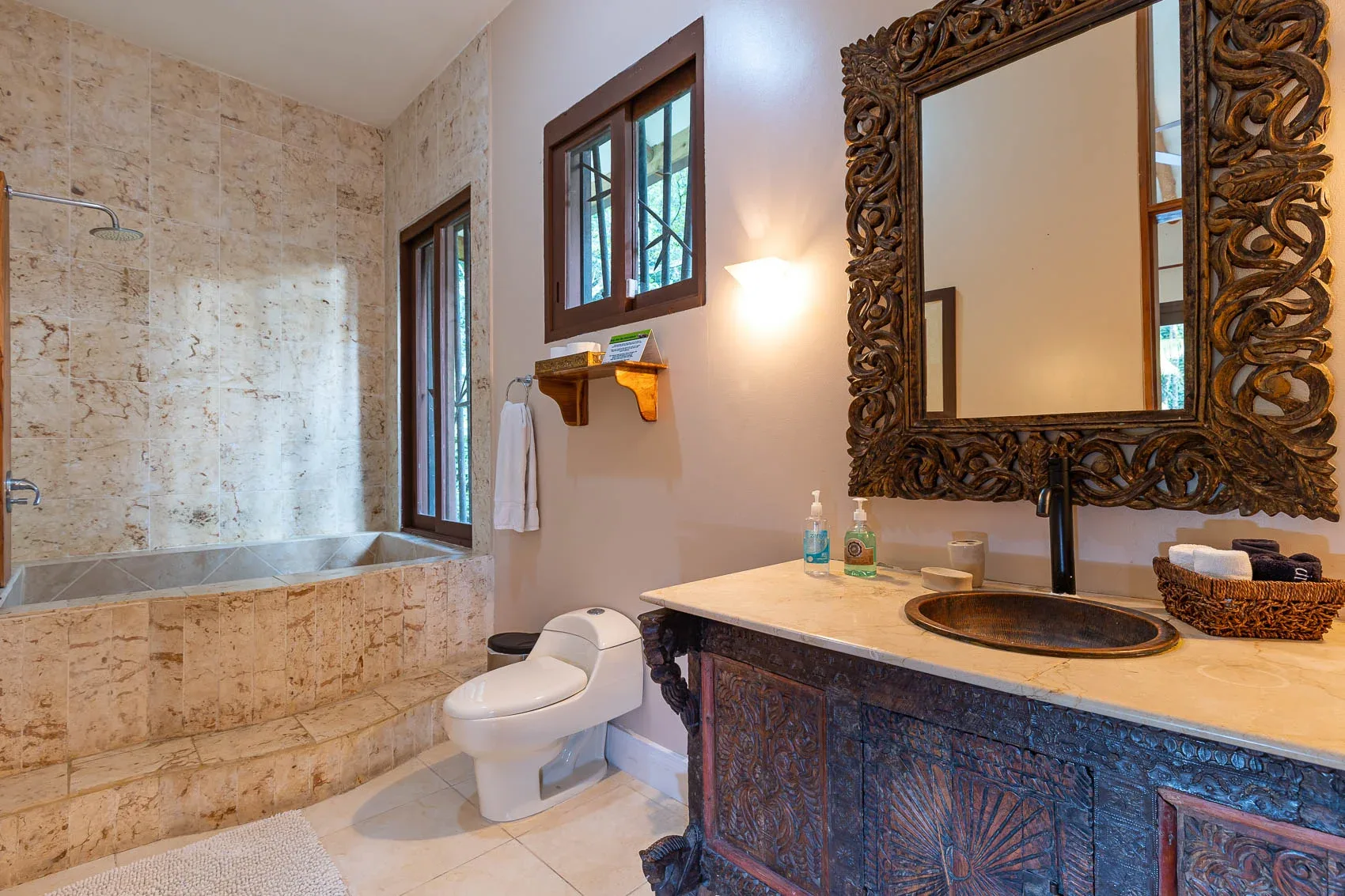 Un baño con bañera revestida de azulejos, inodoro y un espejo ornamentado sobre el tocador.