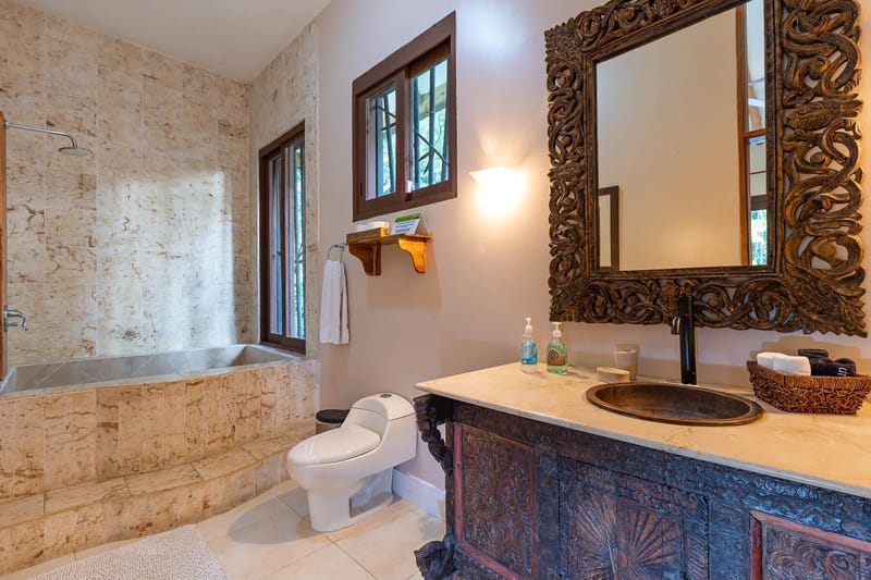 Villa Michelina · 14. This bathroom features a deep, tiled bathtub/shower, a hand-carved vanity with a unique basin under an ornate mirror, and windows offering natural light and views of the lush surroundings.