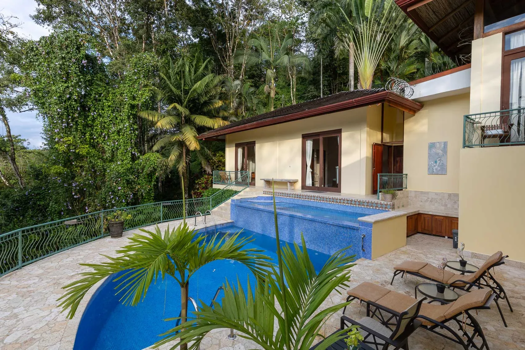 Una piscina y un patio de piedra están junto a un edificio, rodeados de vegetación tropical.