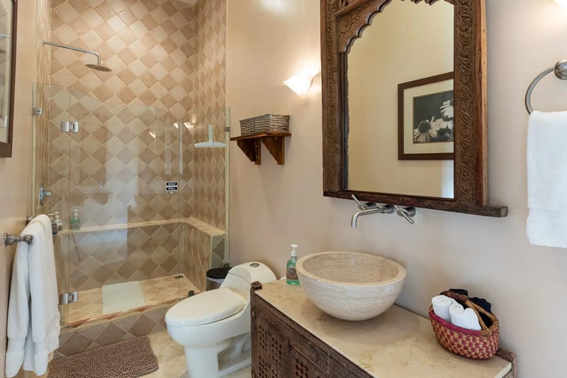 Villa Michelina · 21. A Balinese-inspired bathroom offers a walk-in shower with warm-toned diamond-patterned tiles, a stone vessel sink on a vanity, and an intricately carved wooden mirror.