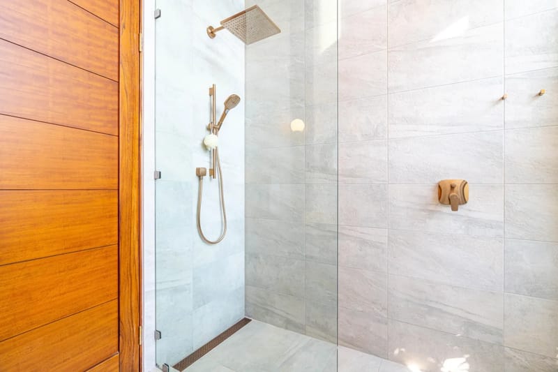 Casa Bella Vina · 14. Este baño cuenta con una ducha de lluvia cerrada con vidrio, revestida con azulejos claros de apariencia pétrea y accesorios de bronce, junto a una puerta de madera de tonos cálidos, todo iluminado con luz ambiental.