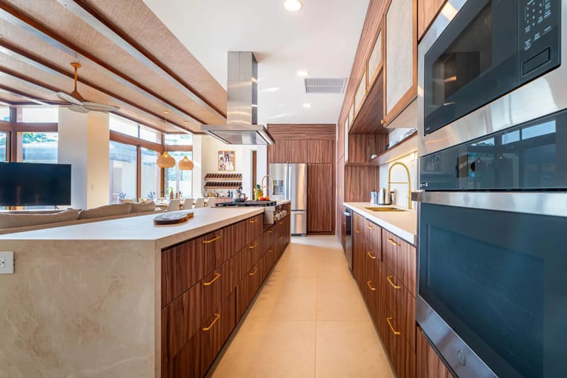 Casa Bella Vina · 8. Esta cocina cuenta con gabinetes de madera oscura, encimeras de piedra y electrodomésticos de acero inoxidable, además de un techo tejido con textura y grandes ventanas que aportan luz natural y vistas al entorno tropical.