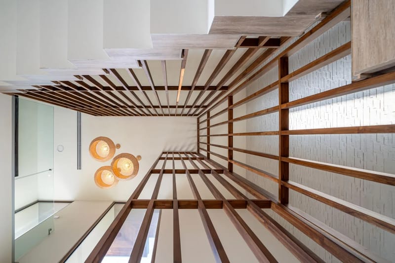 Casa Bella Vina · 10. Una vista hacia arriba muestra un interior de varios niveles con listones de madera oscura en el techo, una escalera blanca con barandal de vidrio, tres lámparas colgantes tejidas y una pared decorativa con textura, destacando los materiales naturales.