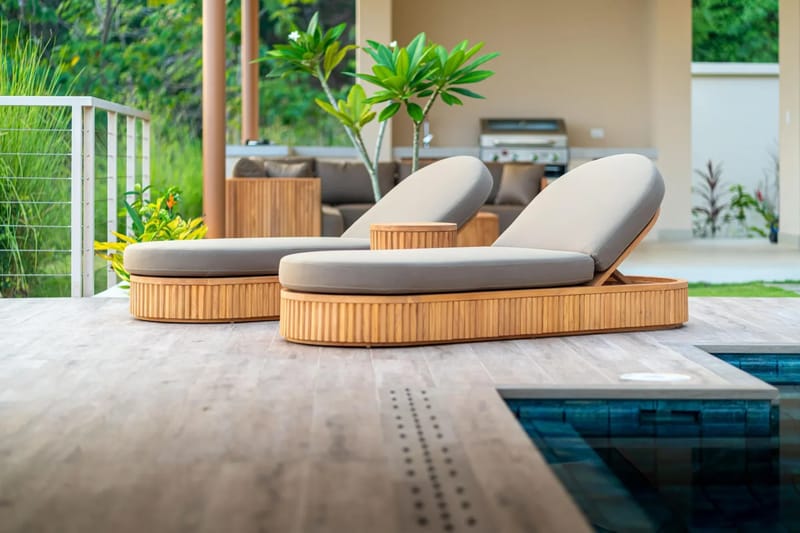 Casa Bella Vina · 20. Una terraza de madera con dos tumbonas se encuentra junto a una piscina infinita, con una cocina exterior cubierta y un sofá visibles entre la vegetación tropical.