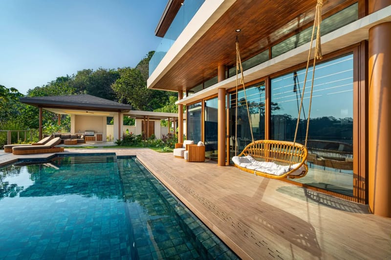 Casa Bella Vina · 18. Una piscina infinita revestida de azulejos de color azul profundo se extiende desde una terraza de madera clara, frente a una villa moderna con puertas de vidrio de piso a techo y vistas a la selva tropical circundante bajo un cielo despejado.