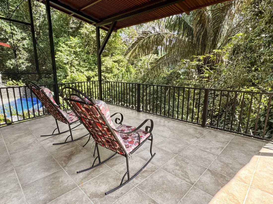 A covered patio has patterned seating and a railing overlooking dense greenery.