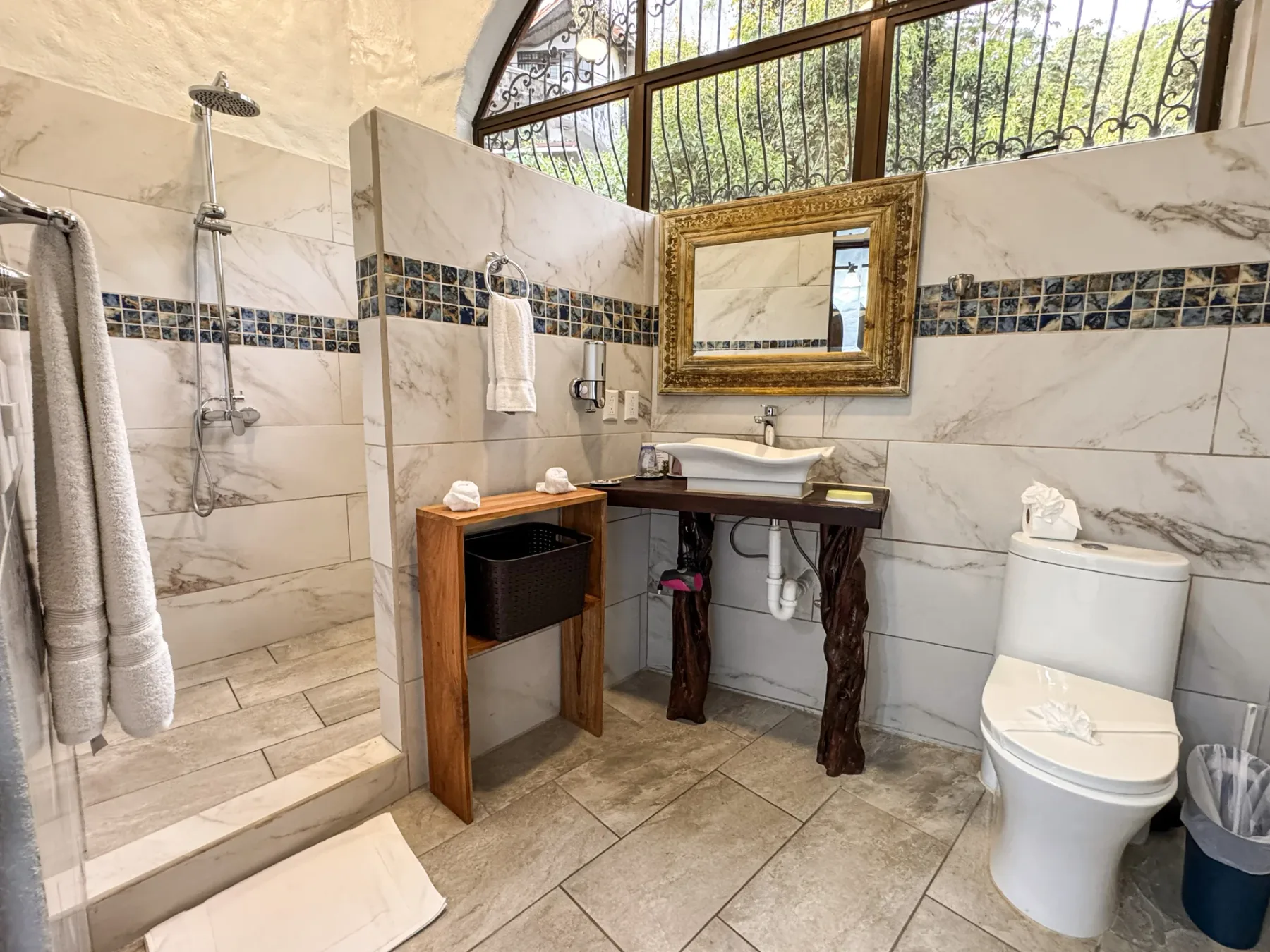 A tiled bathroom with a shower, sink, and toilet beneath an arched window.