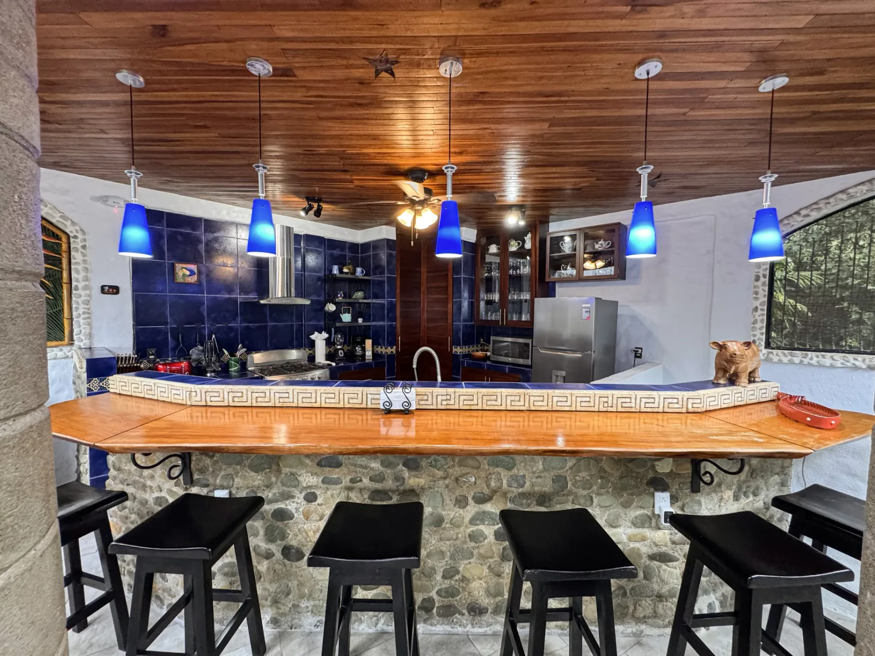 A kitchen bar with dark stools, blue pendant lights, and wood ceilings.