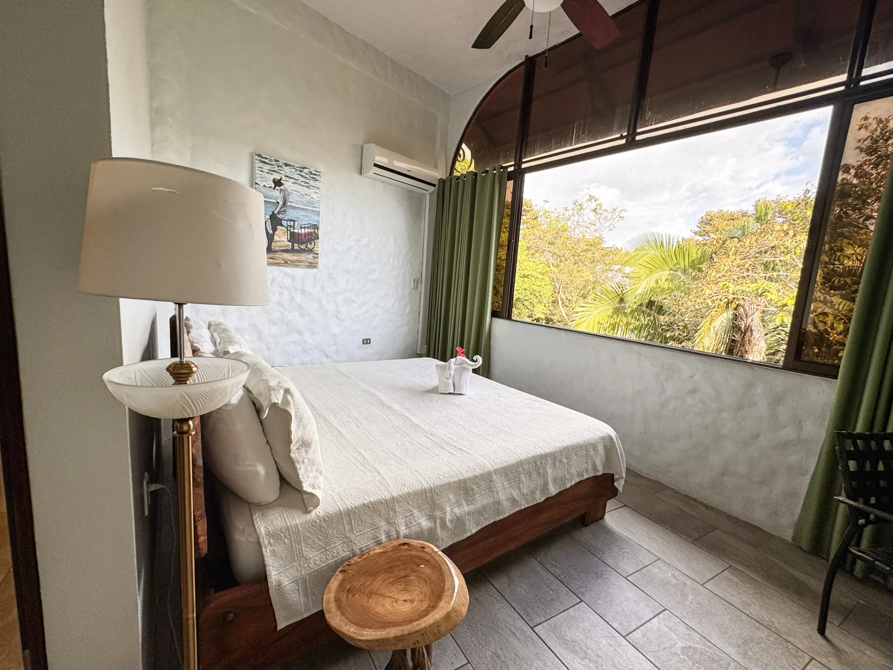 A bedroom with a neatly made bed beside a large arched window overlooking leafy greenery.