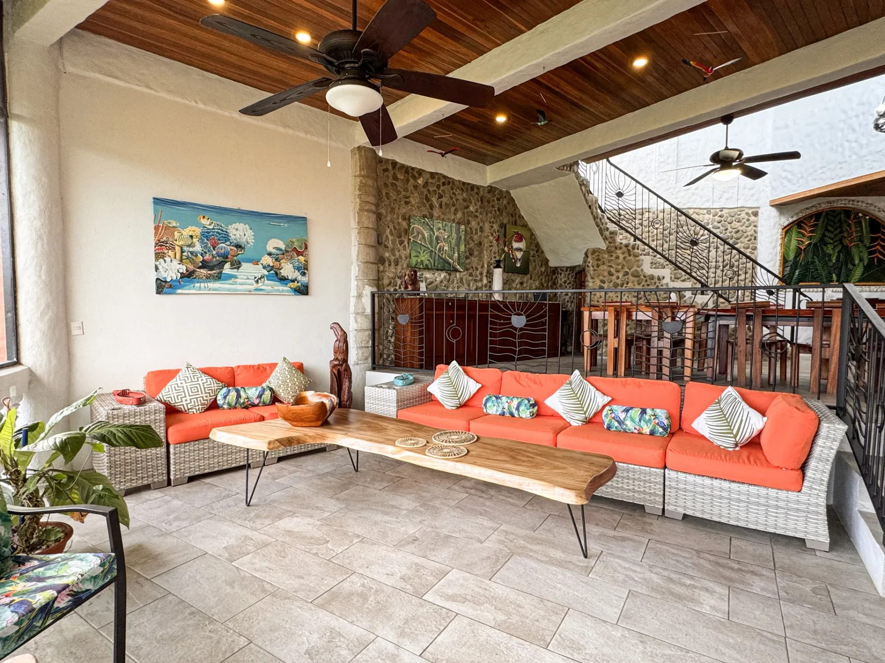 An open-air lounge has woven seating with bright orange cushions beneath a wooden ceiling.