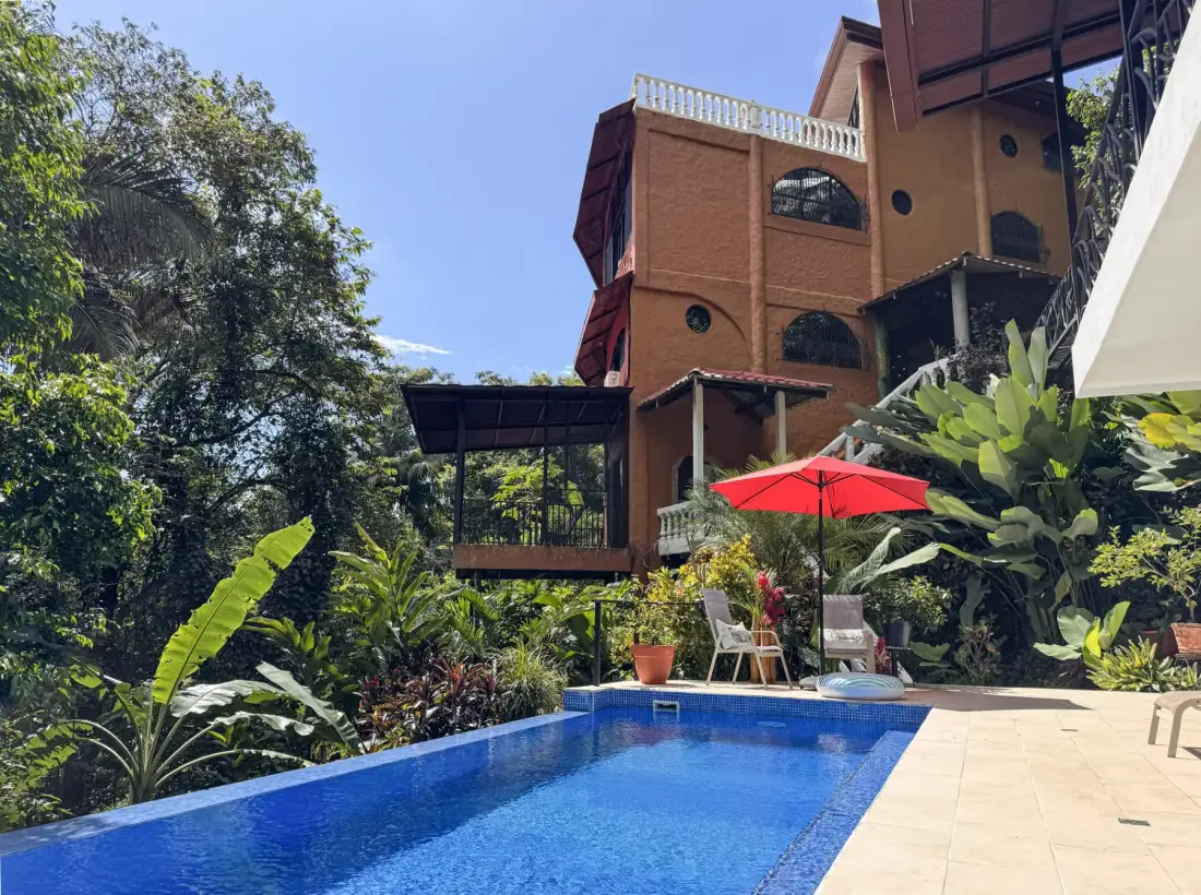 A swimming pool sits beside a multi-story building amid lush tropical plants.