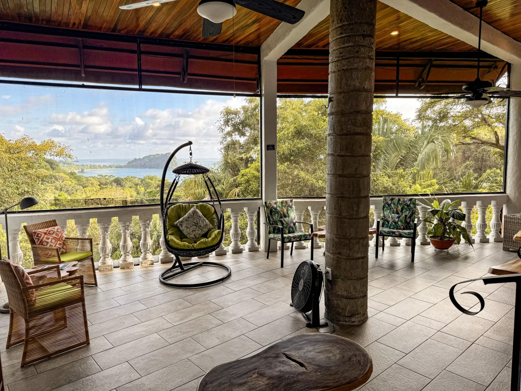 A covered terrace with outdoor seating overlooks greenery and a body of water.