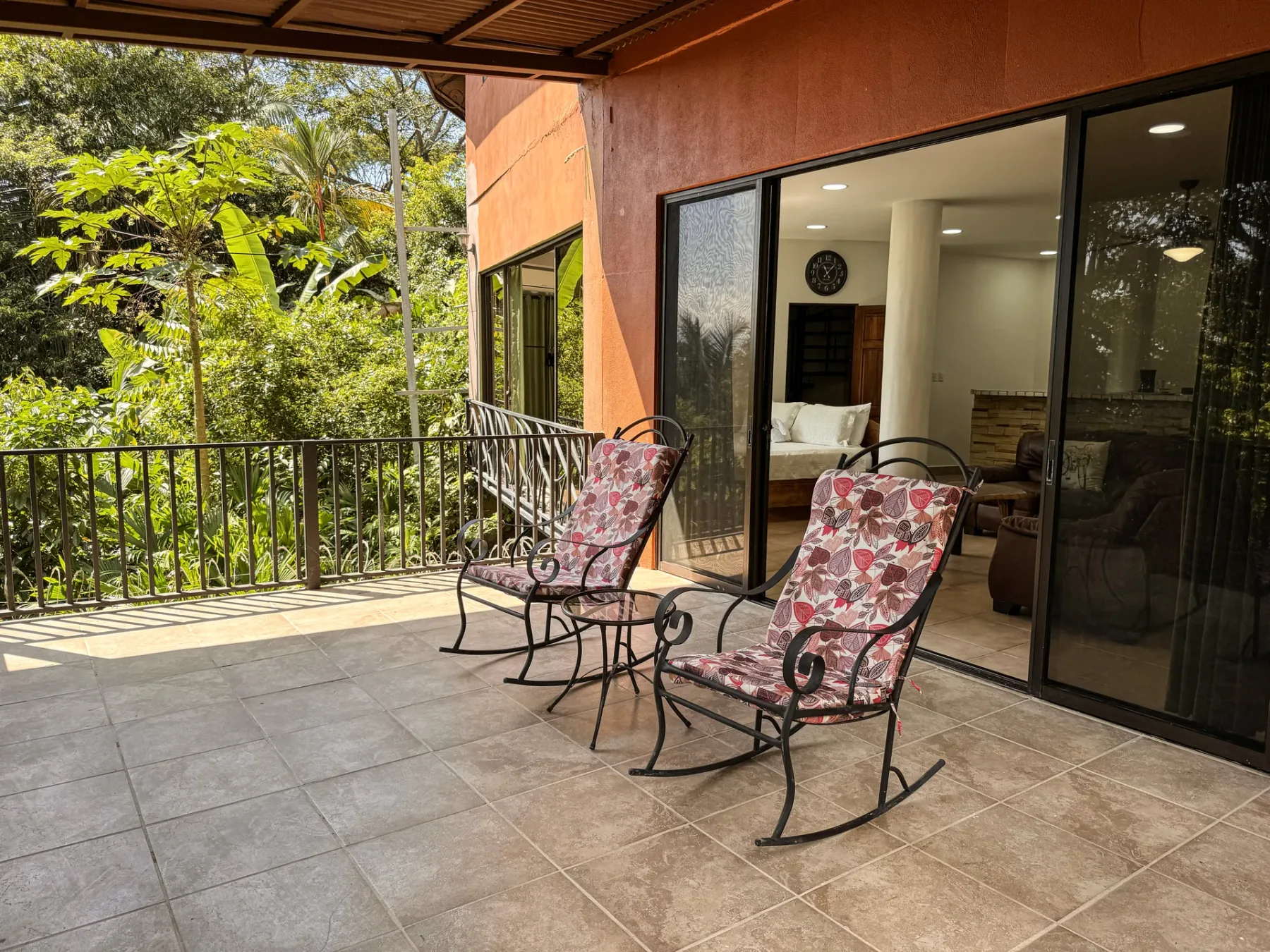 A tiled outdoor terrace has metal railing, two patterned rocking chairs, and views of surrounding trees.