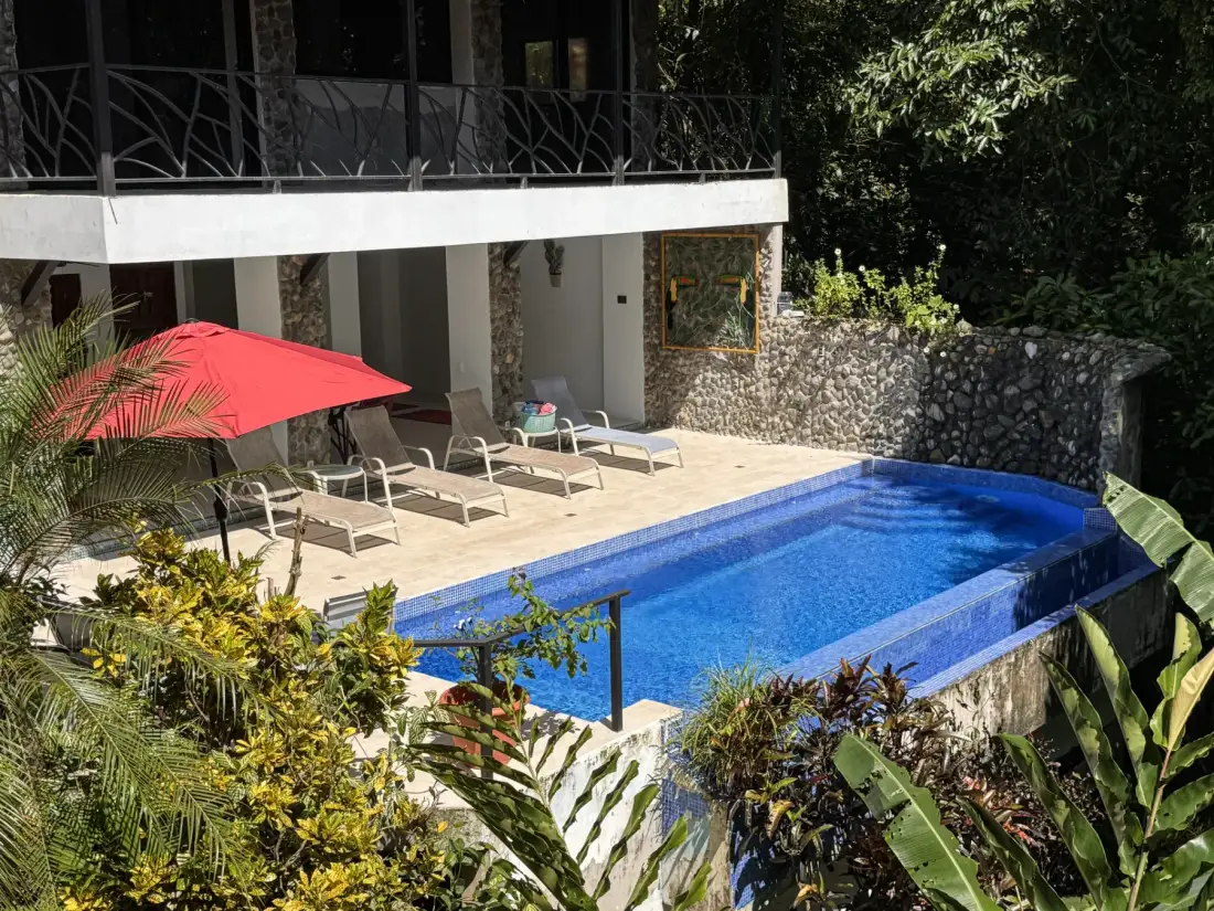 An outdoor swimming pool sits beside a patio with lounge seating and a red umbrella.