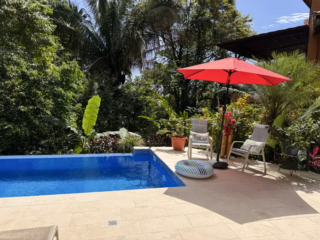 A swimming pool and patio seating sit among lush tropical plants beneath a red umbrella.