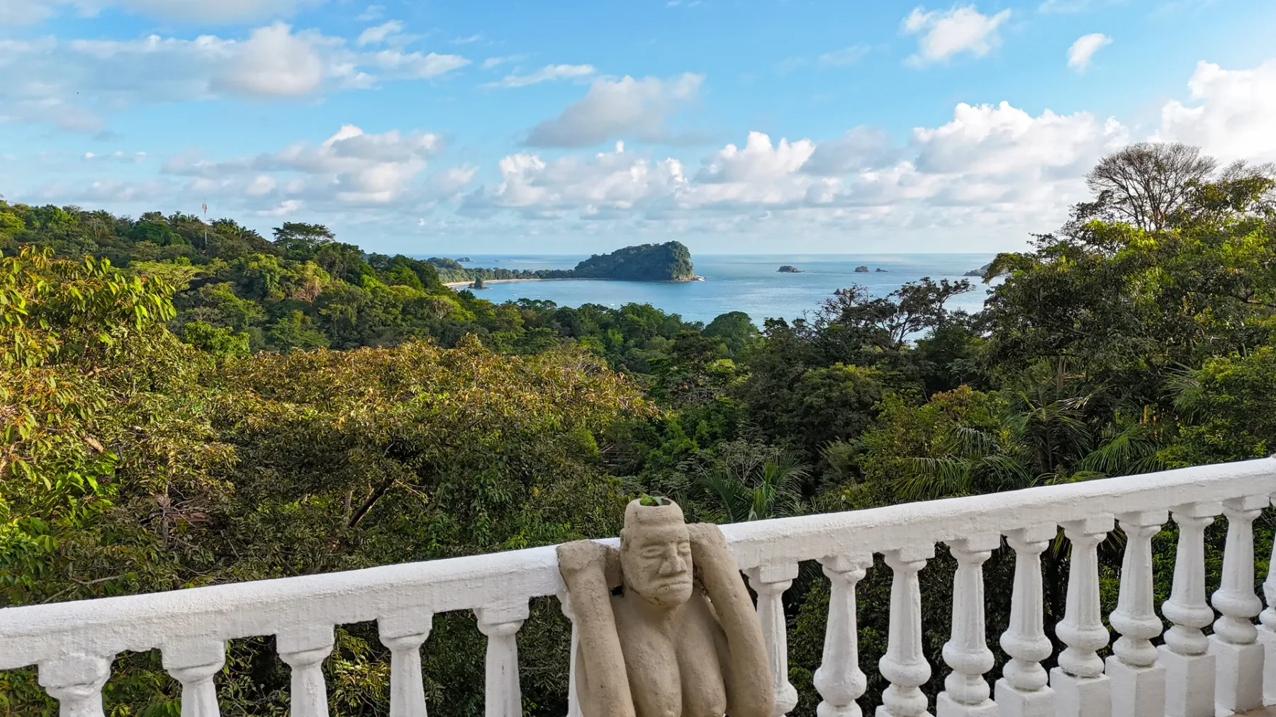 A stone statue stands beside a white balcony railing overlooking a green landscape and water.