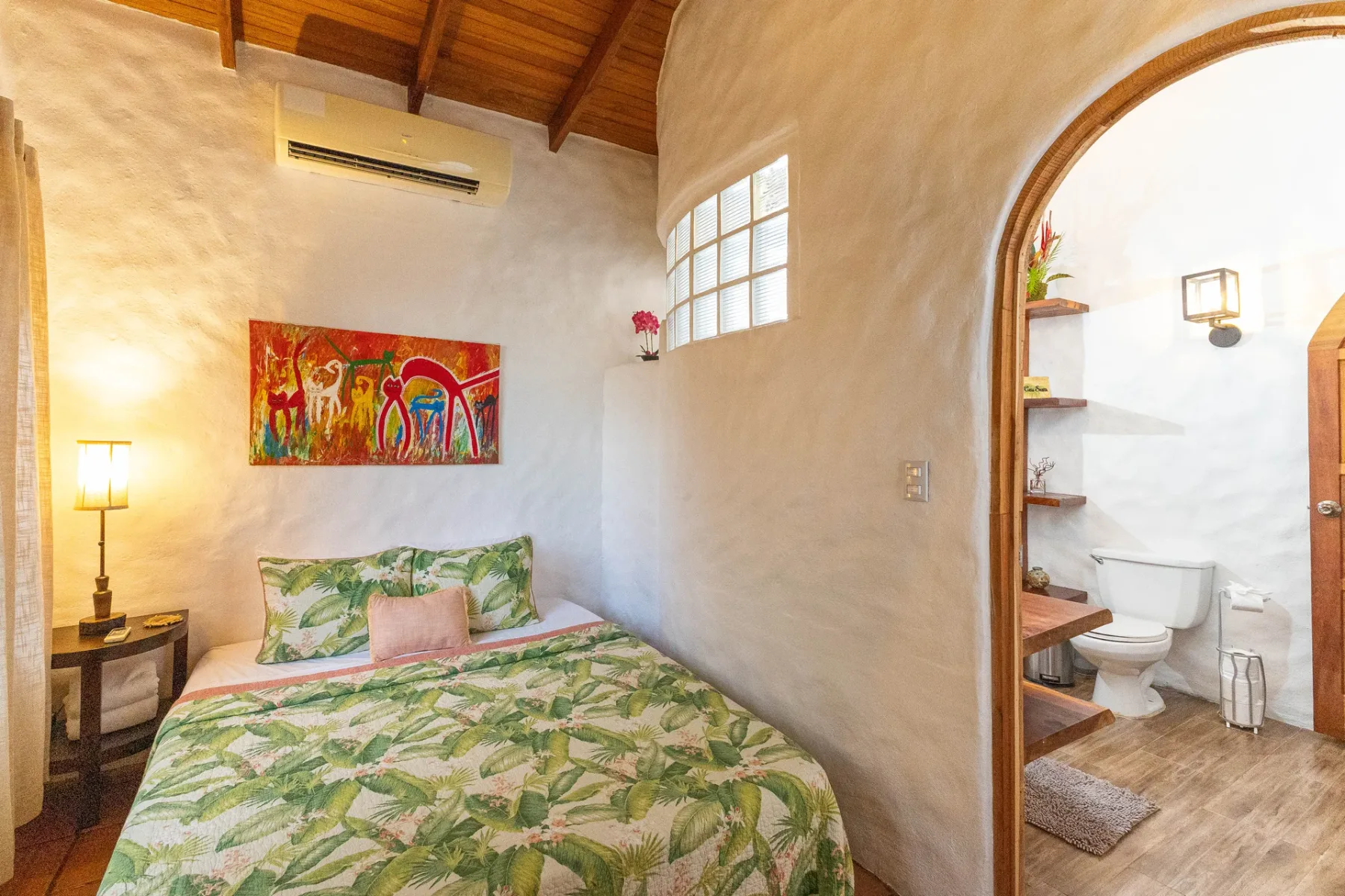 A bedroom with a bed beneath colorful wall art and an arched bathroom doorway.