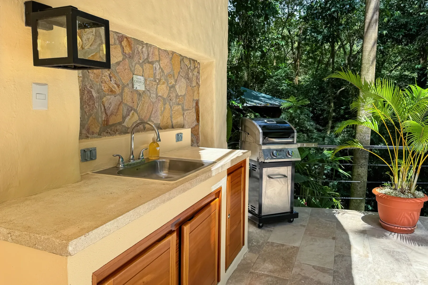 An outdoor kitchen counter with a sink and a grill beside lush greenery.