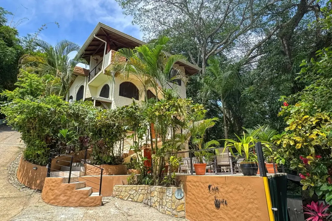 A multi-level house stands behind a lush garden and paved walkway.
