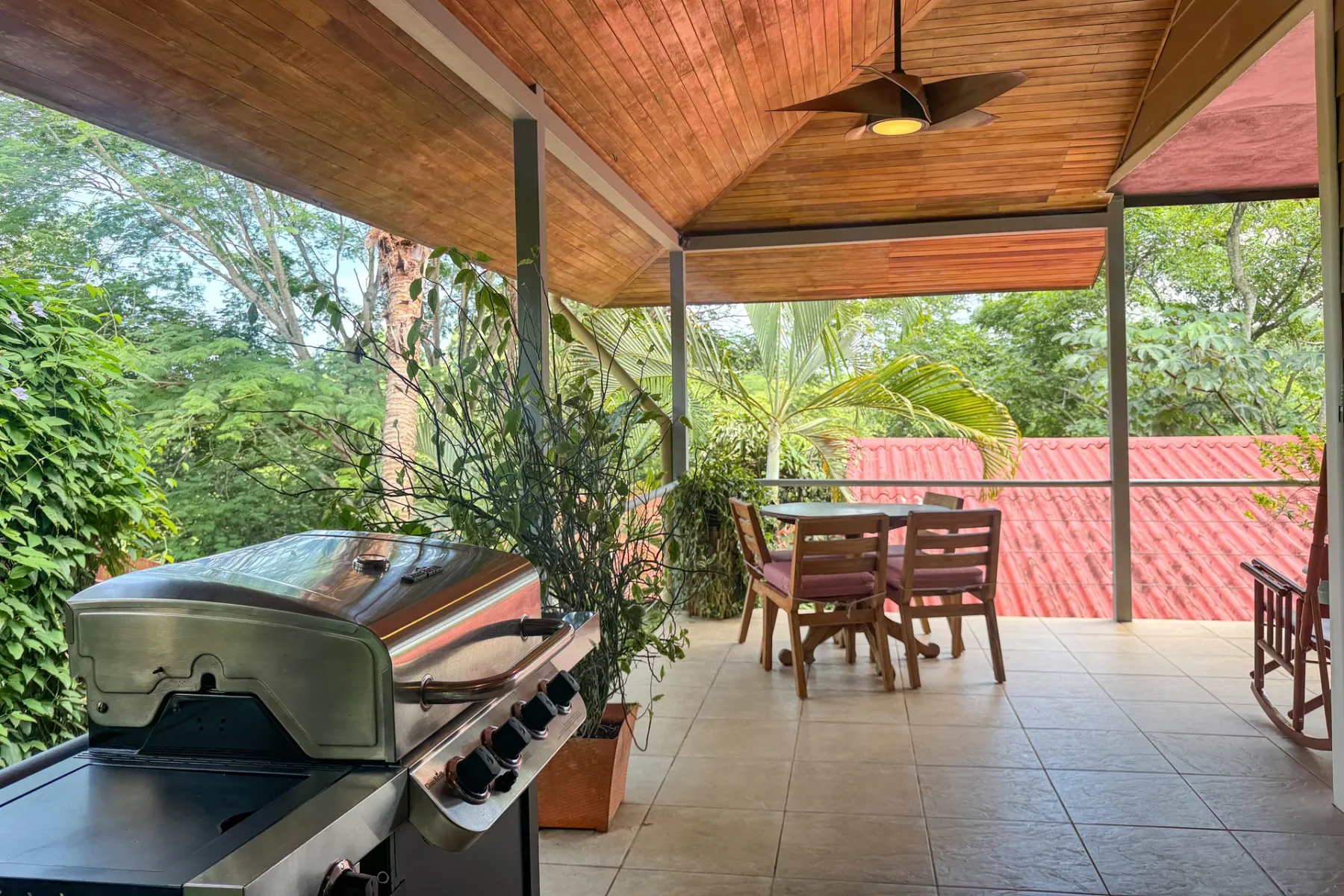 Un comedor exterior cubierto con una mesa y una parrilla da a una zona frondosa.