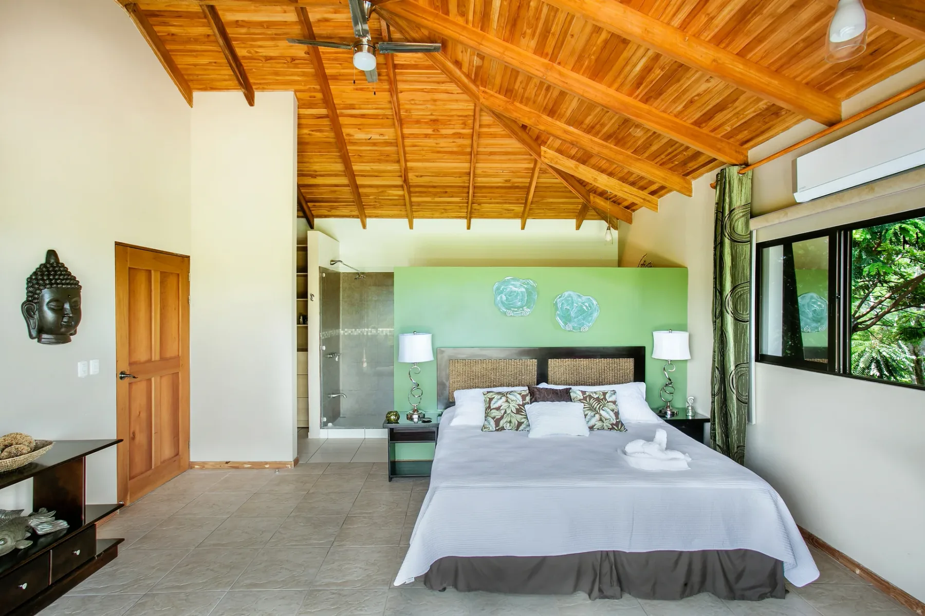 Un dormitorio con una cama bajo un techo alto de madera y ventanas que dan a la vegetación.