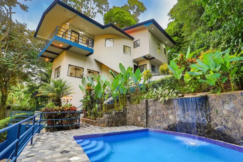 Casa Elena · 7. The multi-level contemporary villa with blue trim overlooks a private pool featuring a waterfall, surrounded by dense rainforest vegetation.