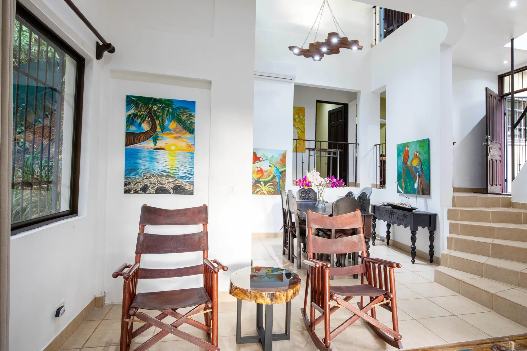 A bright living area with wooden seating, colorful paintings, and a staircase.