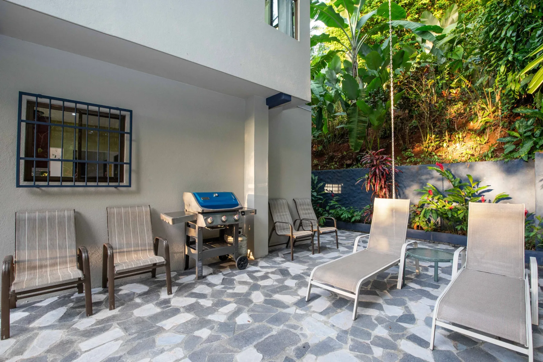 A paved outdoor patio has lounge seating, a grill, and dense greenery.
