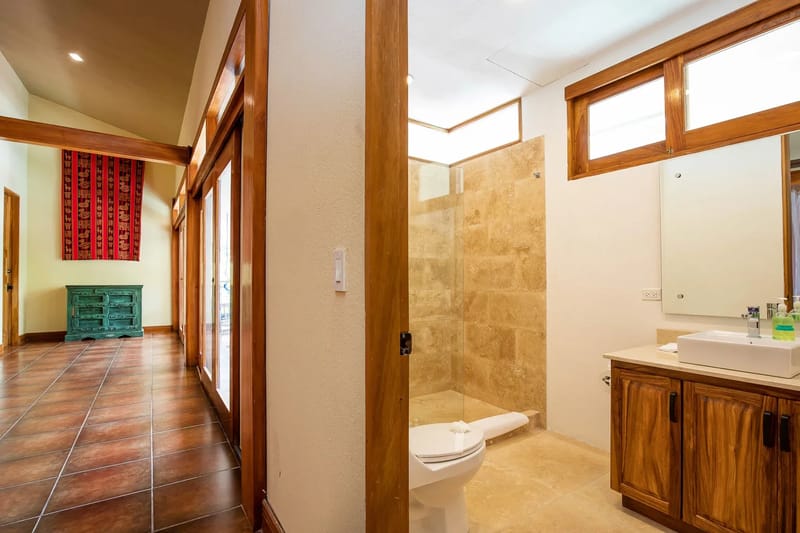 The Rainforest Estate · 16. The bathroom features a spacious tiled walk-in shower and a wooden vanity, illuminated by natural light from transom windows, and connects to a hallway with large glass doors.
