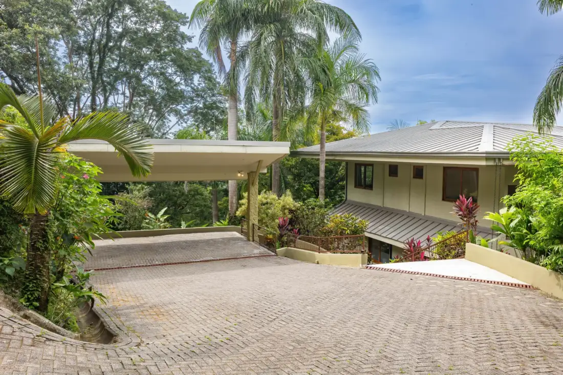Una entrada pavimentada conduce a un área de estacionamiento cubierta junto a un edificio rodeado de vegetación tropical.