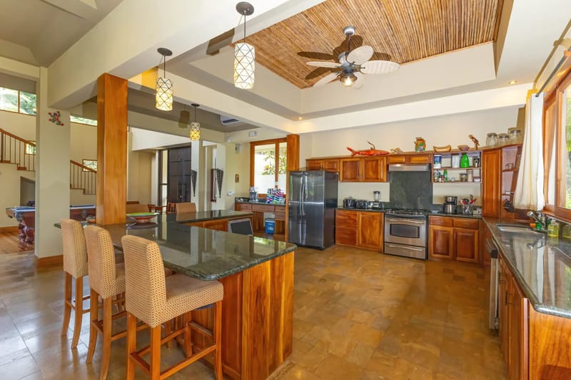 The Rainforest Estate · 7. This spacious kitchen features warm wood cabinetry, dark granite countertops, an island with seating, a bamboo-paneled ceiling, and a large window offering natural light and views of tropical foliage.