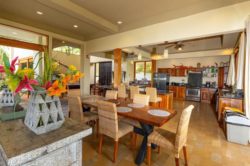 The Rainforest Estate · 4. An expansive, fully equipped kitchen with a central island and breakfast bar flows directly into a spacious dining area with a large wooden table, all illuminated by wide openings to the outdoors.