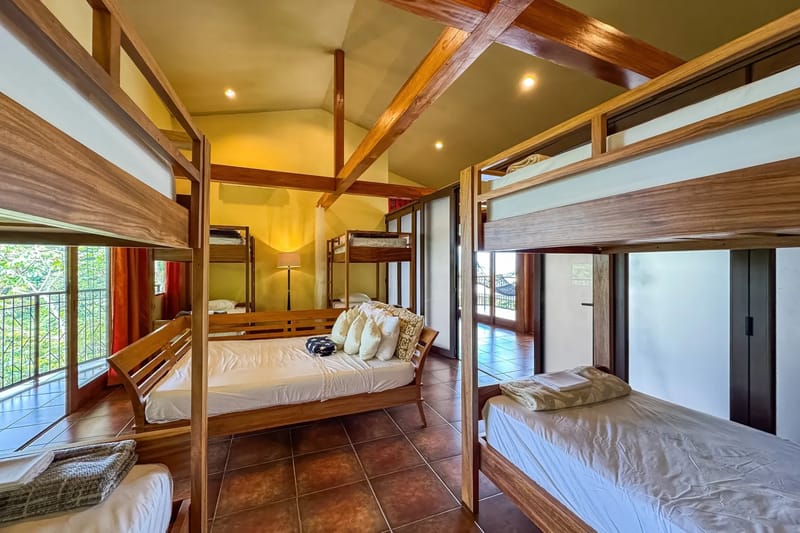 The Rainforest Estate · 15. A bright, shared bedroom with high vaulted ceilings, exposed wood beams, multiple bunk beds, and a double bed, provides views of the rainforest and access to an adjacent outdoor space.