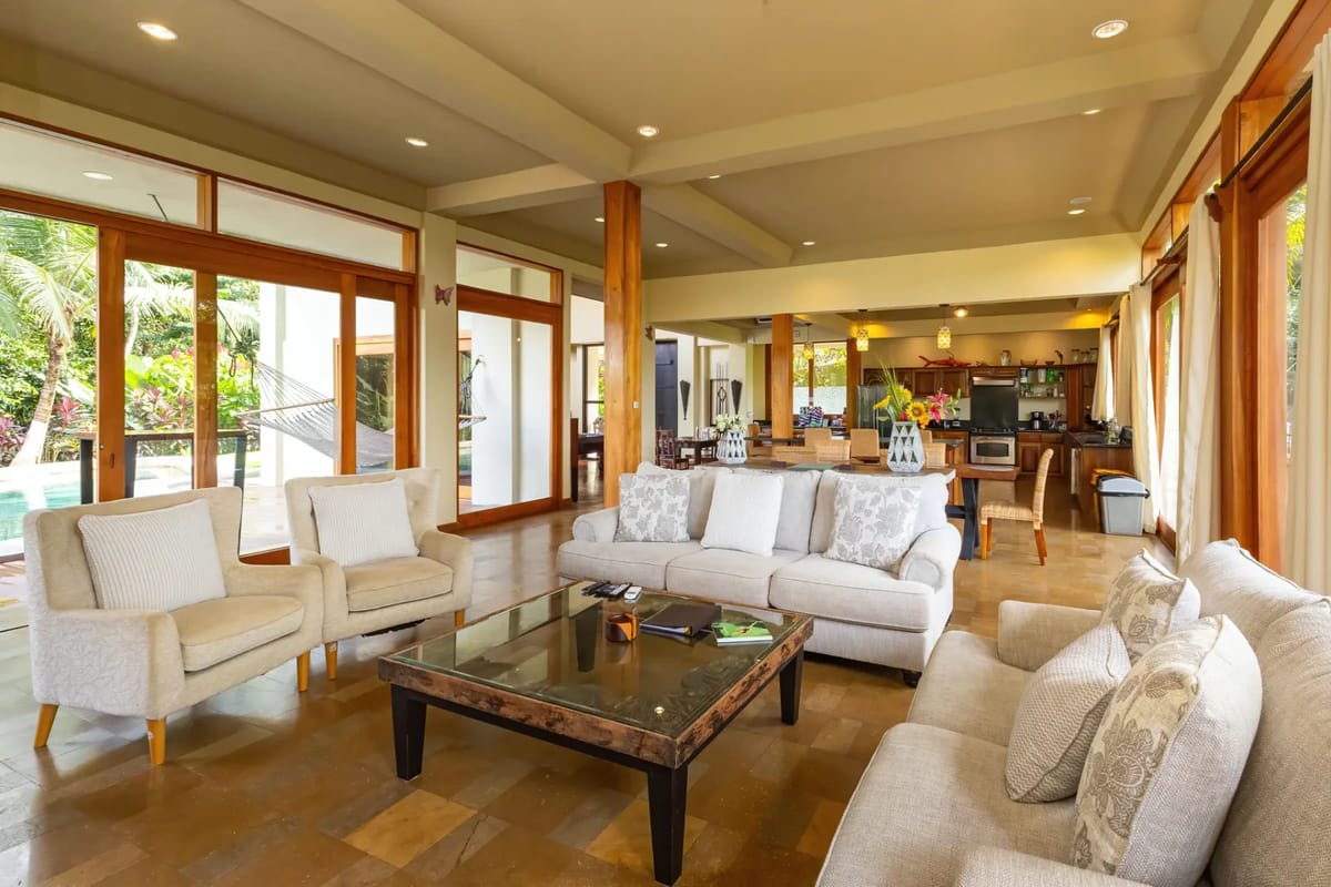 The Rainforest Estate · 3. A bright and expansive living room with multiple seating areas flows into an open kitchen and dining space, connected by large sliding glass doors to a pool deck and tropical views.
