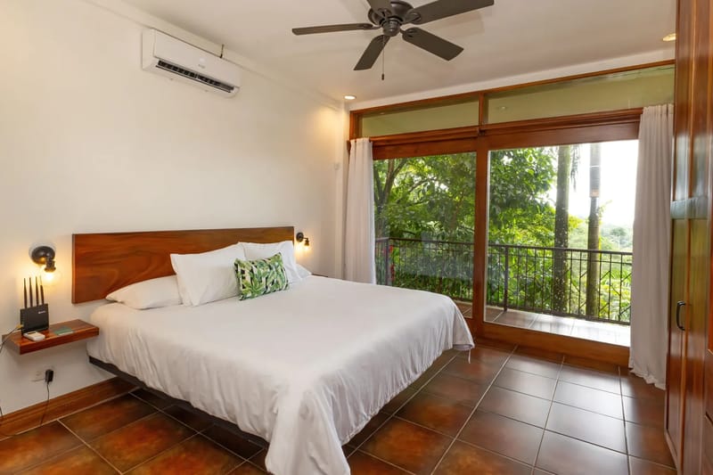 The Rainforest Estate · 18. A serene king bedroom features a ceiling fan and air conditioning, with a large wooden-framed sliding glass door providing natural light and access to a private balcony overlooking the rainforest.