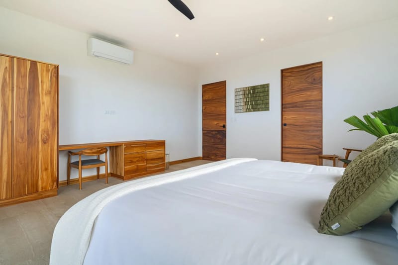 Villa del Cielo · 15. This serene bedroom features a large bed, warm-toned wooden wardrobe, desk with a chair, and two matching wooden doors set against bright white walls, complemented by a ceiling fan and air conditioning.